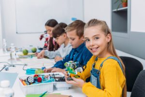 Vier Kinder sitzen an einem Tische und bauen technische Geräte aus Lego auf. Ein Mädchen im Vordergrund lächelt in die Kamera